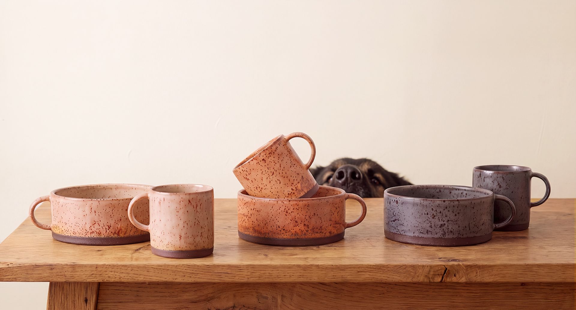Three matching speckled ceramic mug and dog bowl pairs in cream, orange, and purple on a wooden table, with a dog peeking over the middle pair.